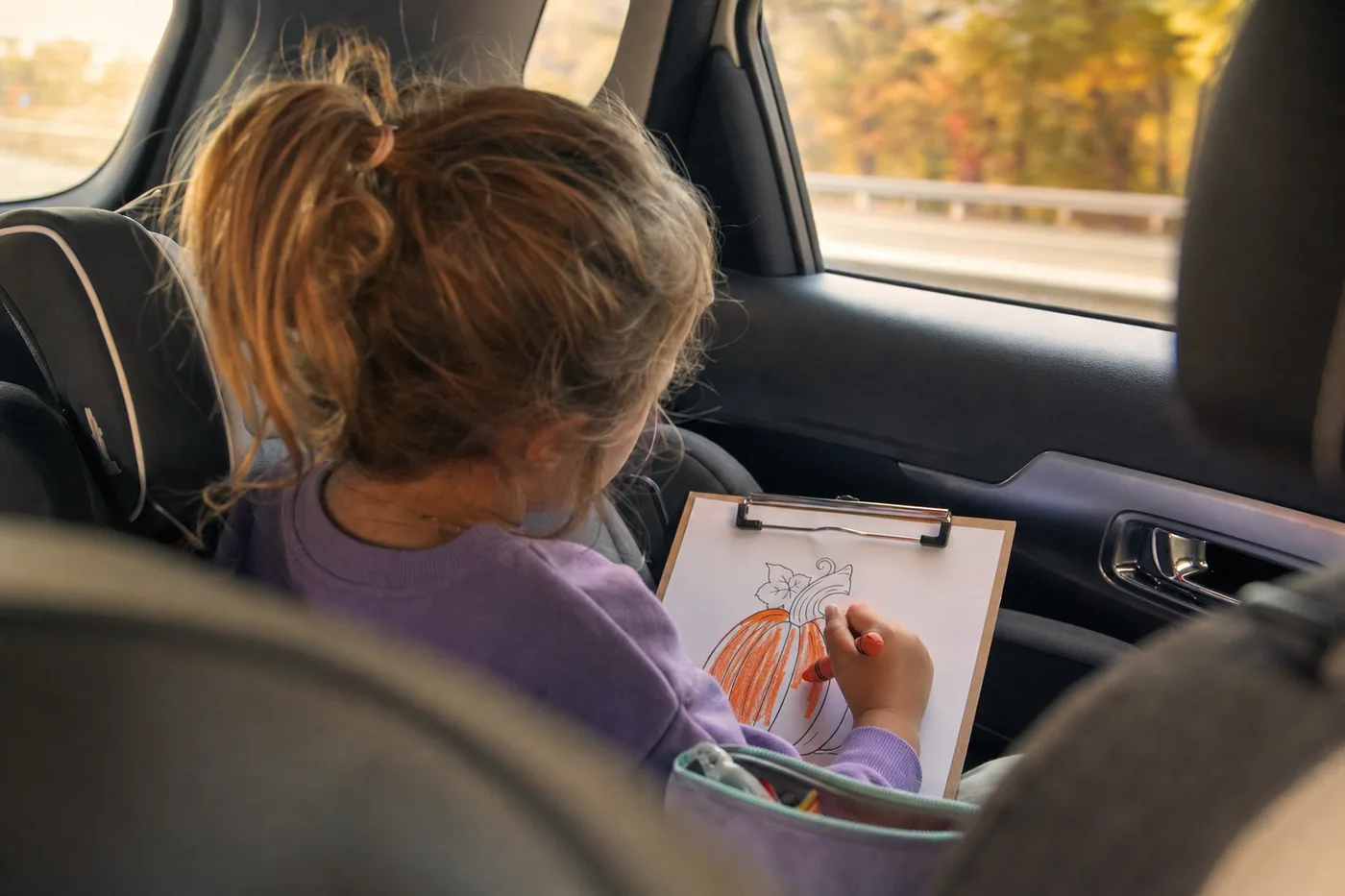 A child coloring a pumpkin page on a clipboard with a crayon in the back seat of a car