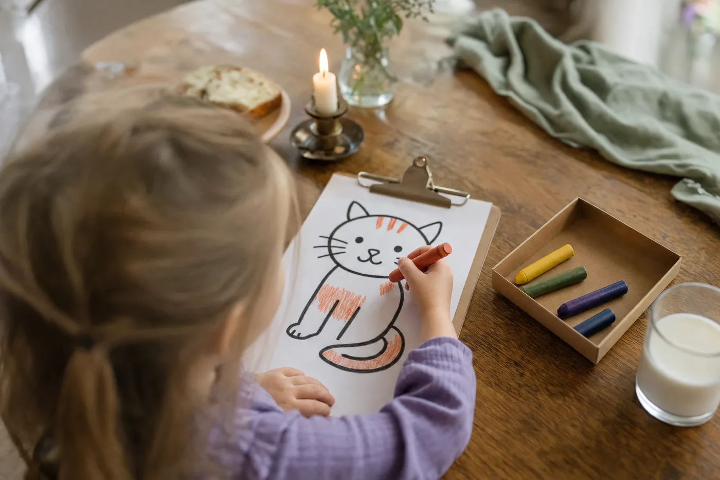 A child coloring a cat page on a clipboard with a chunky crayon at a table, with a box lid holding crayons